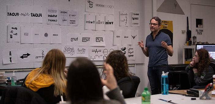 A BSU professor speaks at the front of a graphic design class beside a wall covered with graphic depictions of words