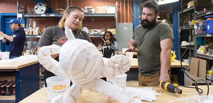 BSU student 和 woodworking professor discuss the student's project, a wooden framed octopus covered in white fabric, while 2 other students work in the background